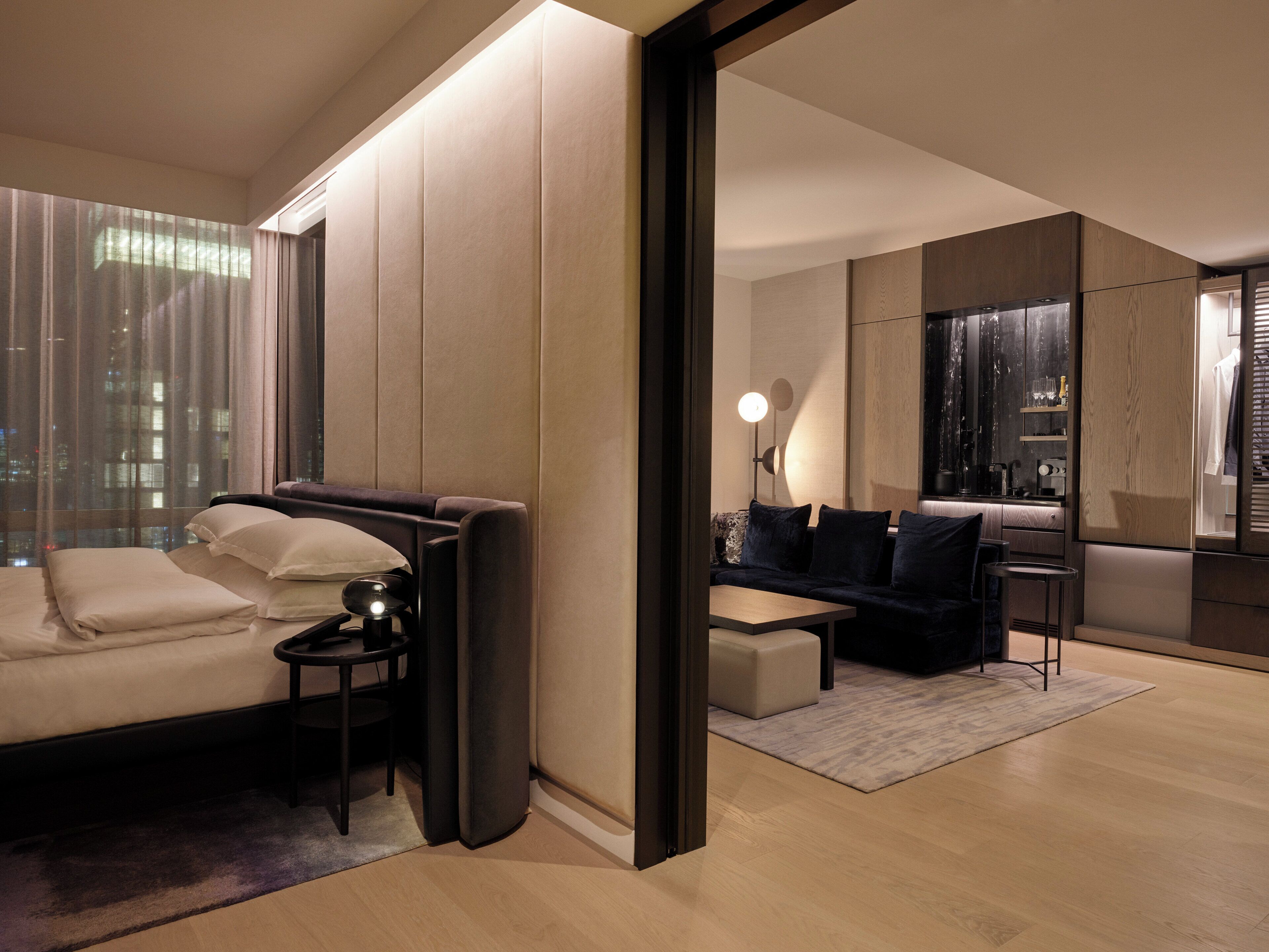 Habitación de hotel moderna con sofá, una mesita, una silla, un espejo y vistas panorámicas de la ciudad a través de amplias ventanas.