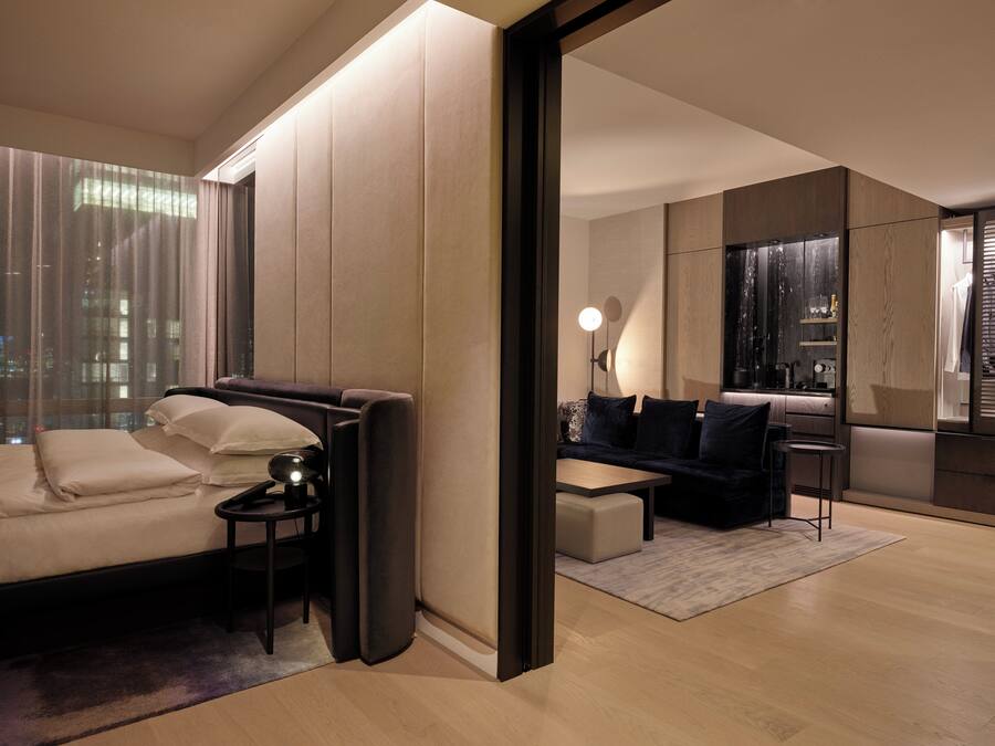 Habitación de hotel moderna con sofá, una mesita, una silla, un espejo y vistas panorámicas de la ciudad a través de amplias ventanas.