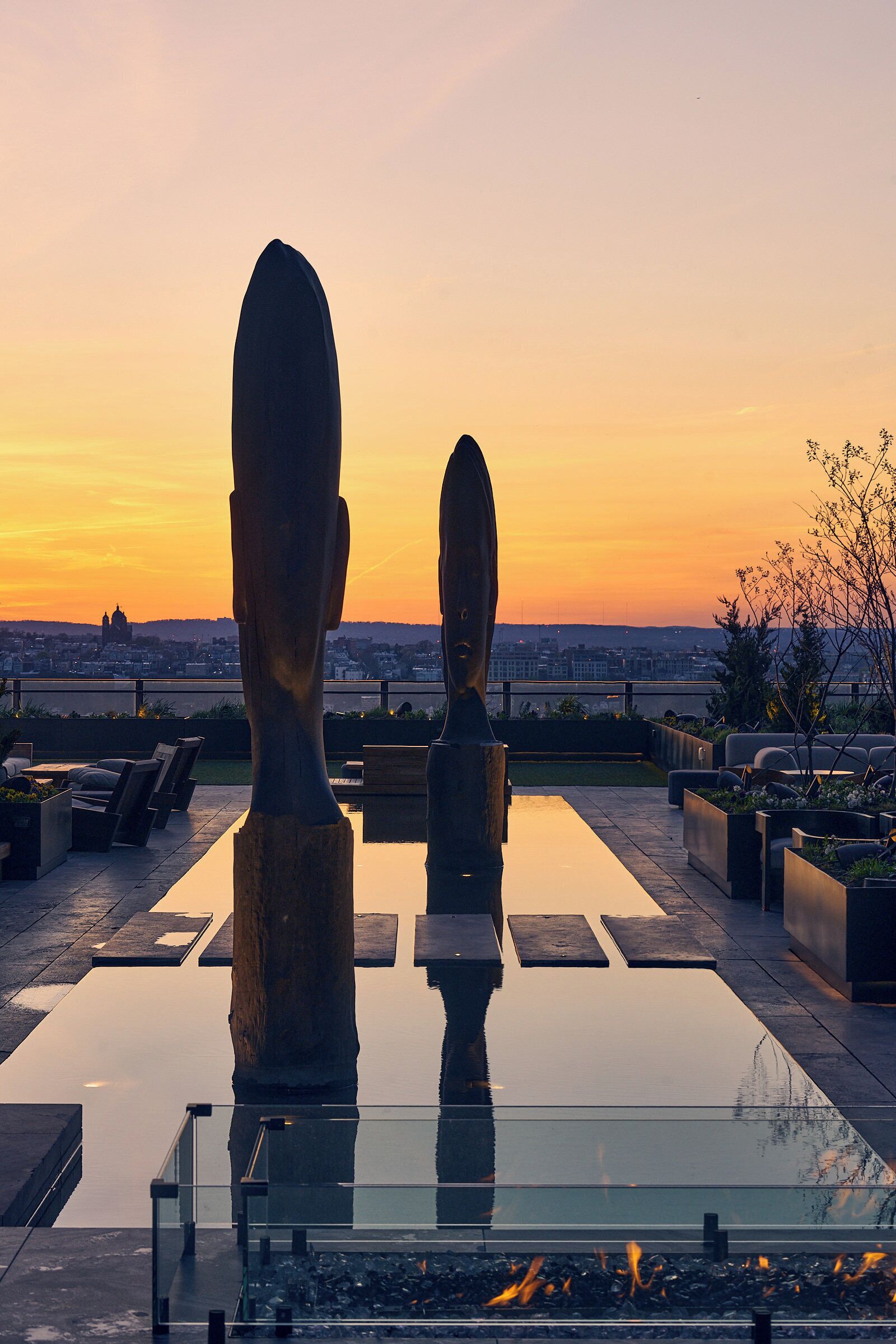 A rooftop terrace with a glass table, two stone sculptures, and a cityscape view at sunset.