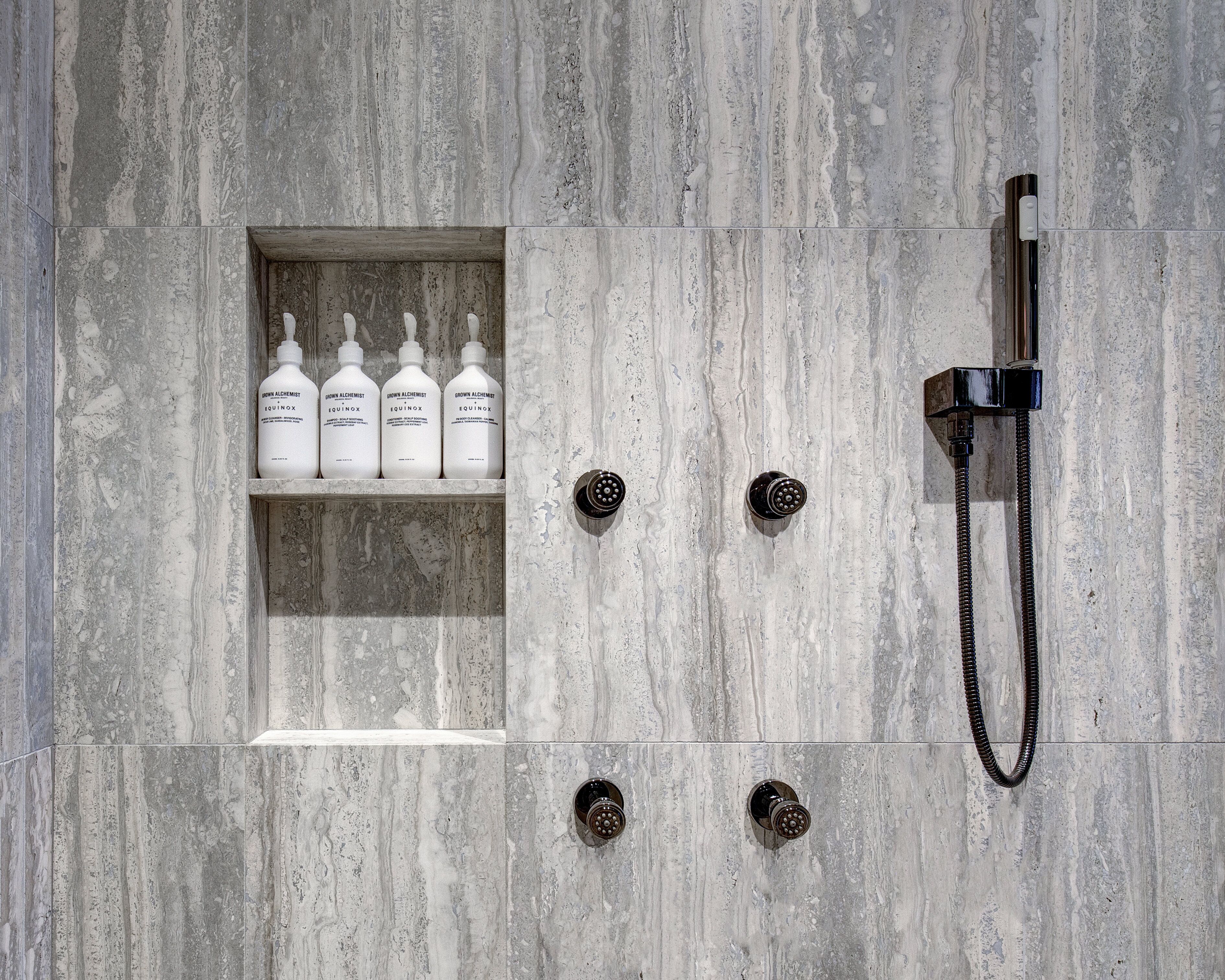 A bathroom with a marble wall, a shelf with bottles, and a showerhead.