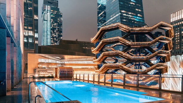 Piscina com placa de 'NÃO MERGULHAR', cercada por arquitetura moderna e paisagem urbana à noite.