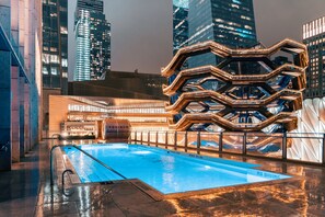 Une piscine avec un panneau indiquant « INTERDIT DE PLONGER », entourée d’une architecture moderne et offrant une vue sur le paysage urbain illuminé la nuit.