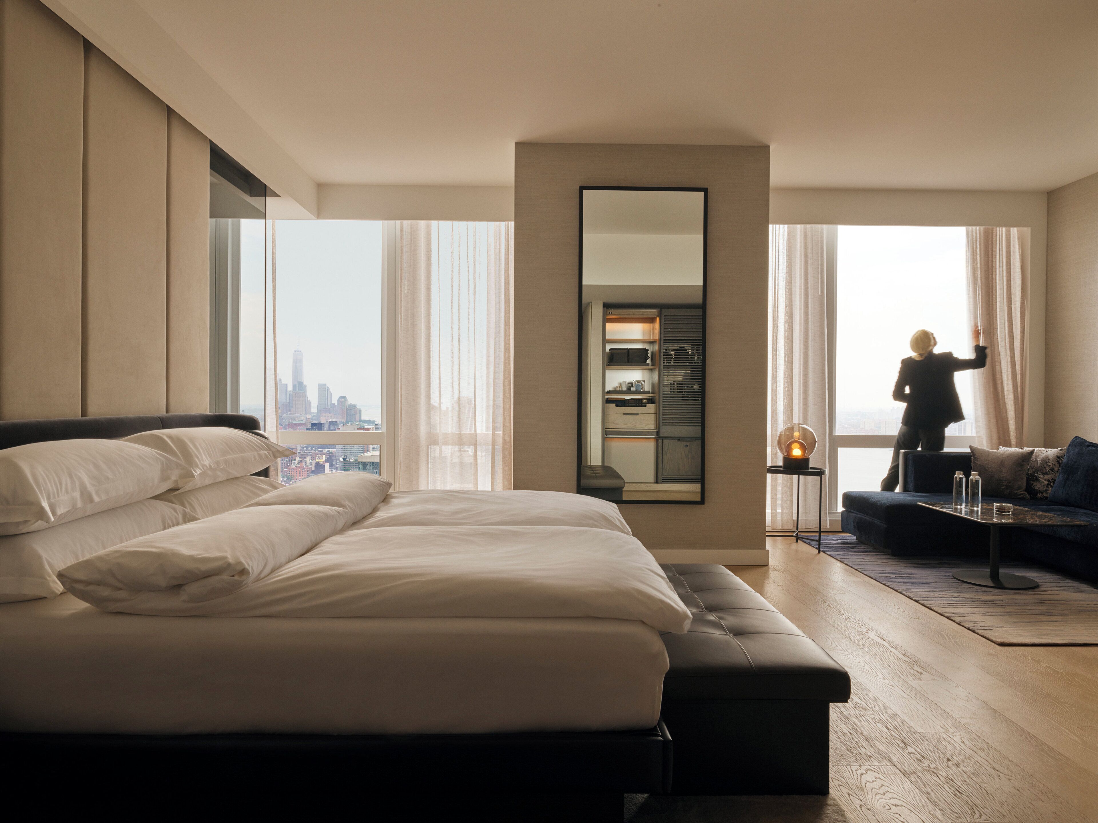 A modern hotel room with a large bed, a seating area, a wardrobe, and a view of the cityscape.