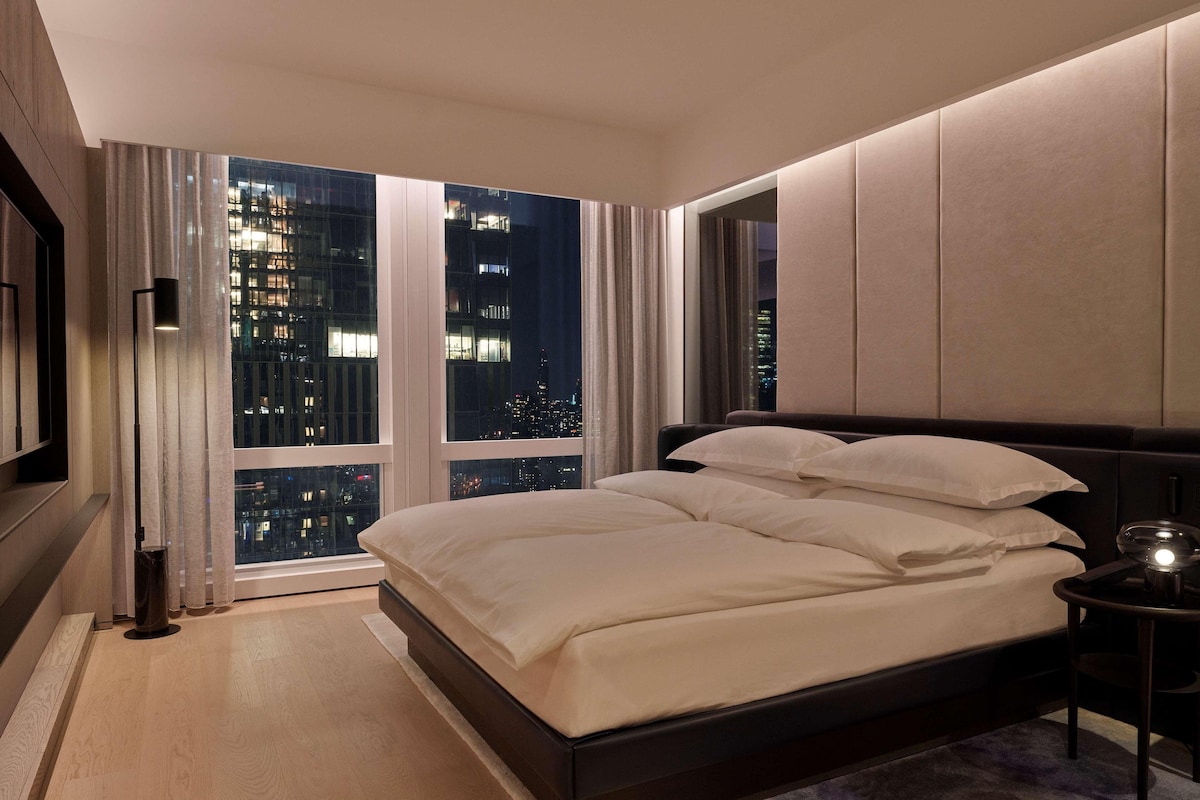 Een moderne hotelkamer met een groot bed, een nachtkastje, een lamp en uitzicht op de stad door ramen van de vloer tot aan het plafond.
