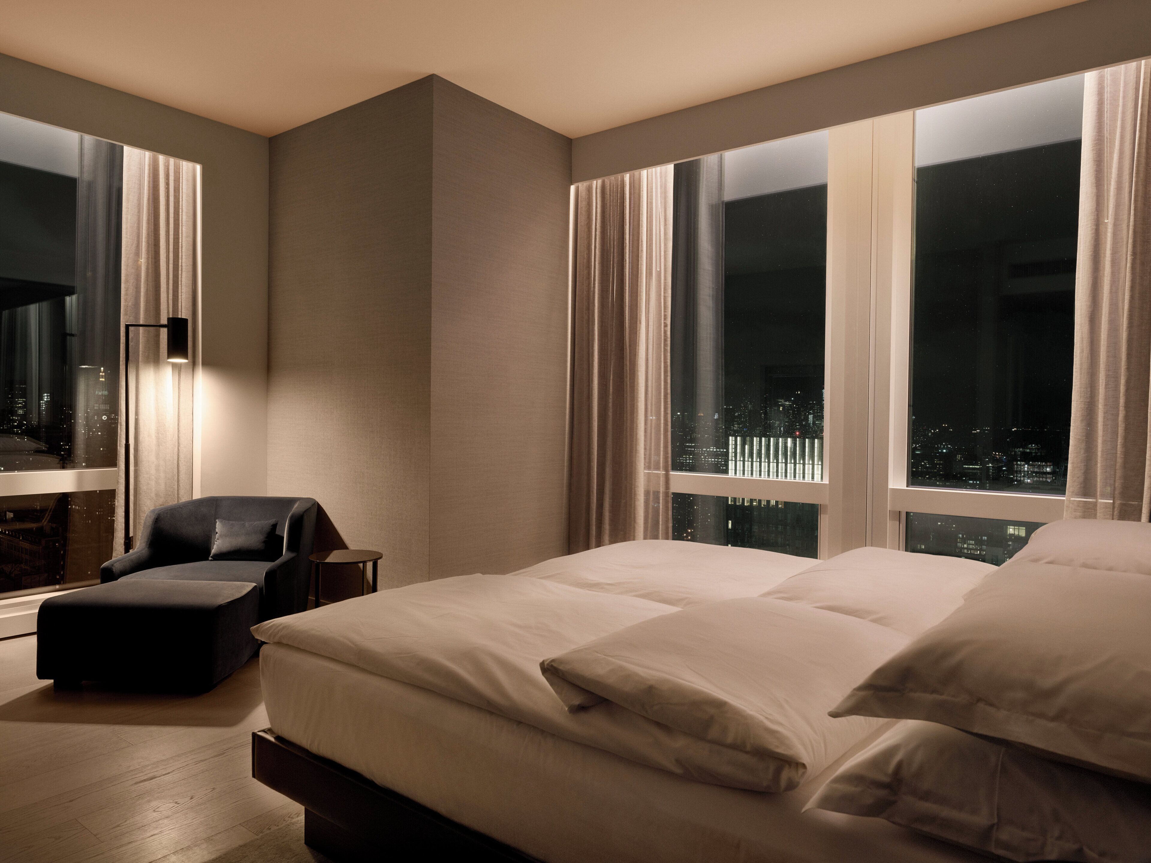 A modern hotel room with a large bed, a seating area, and a city view through the windows.