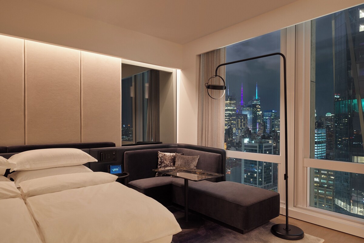 Une chambre d’hôtel moderne dotée d’un grand lit, d’un coin salon et offrant une vue sur le paysage urbain la nuit.
