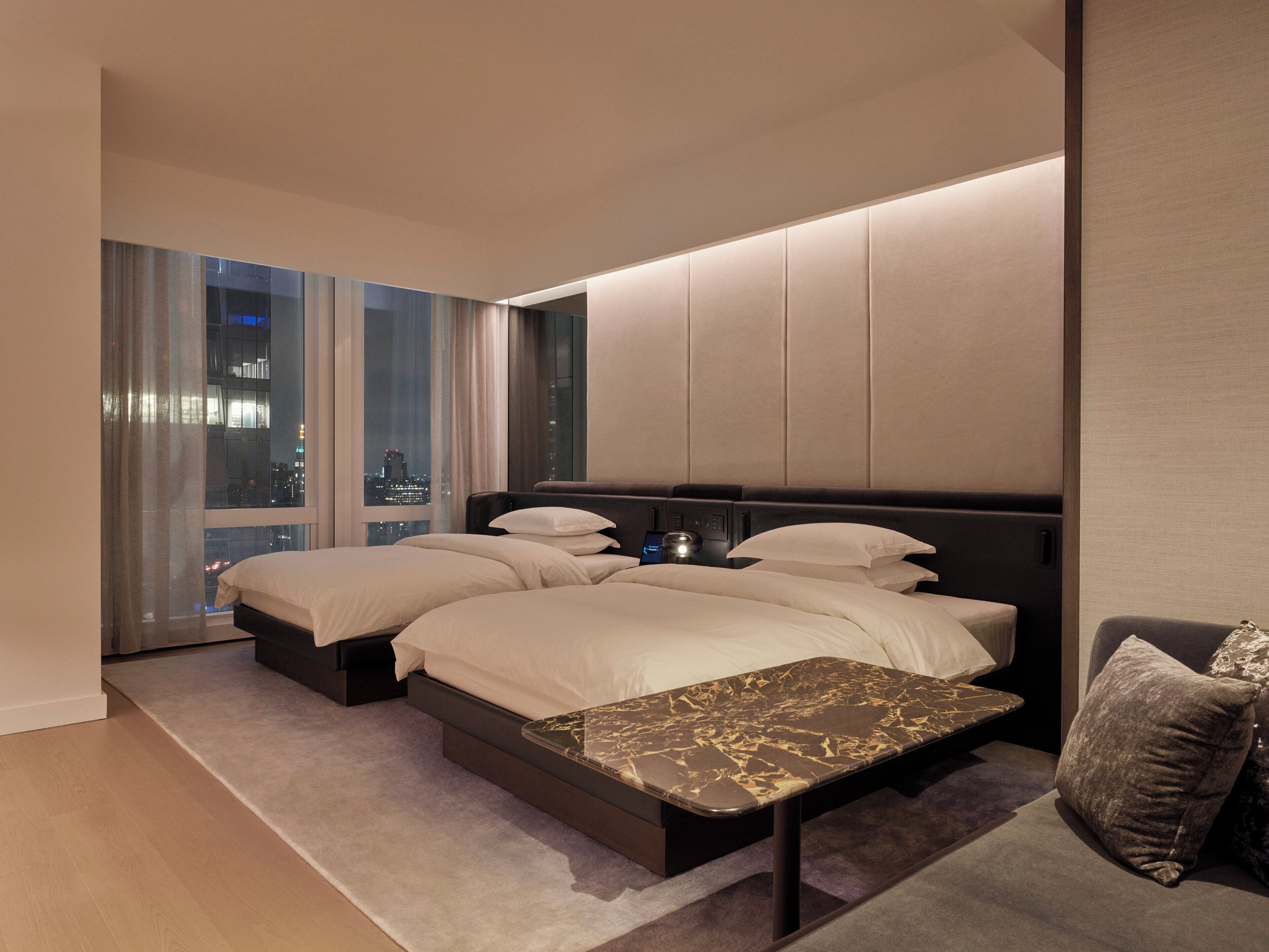 A modern bedroom with a large bed, bedside tables, a nightstand, and a city view.