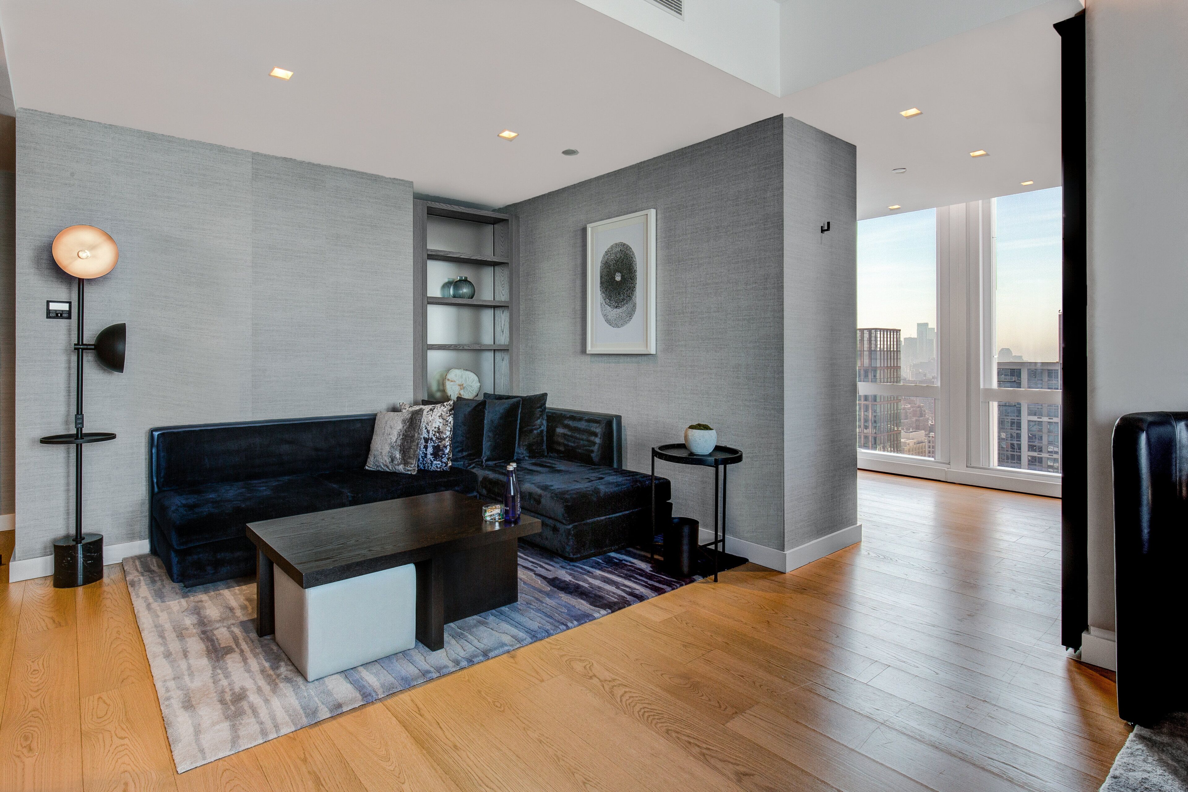 Un salon moderne avec un canapé foncé, une table basse, un lampadaire et une vue sur la ville à travers de grandes fenêtres.