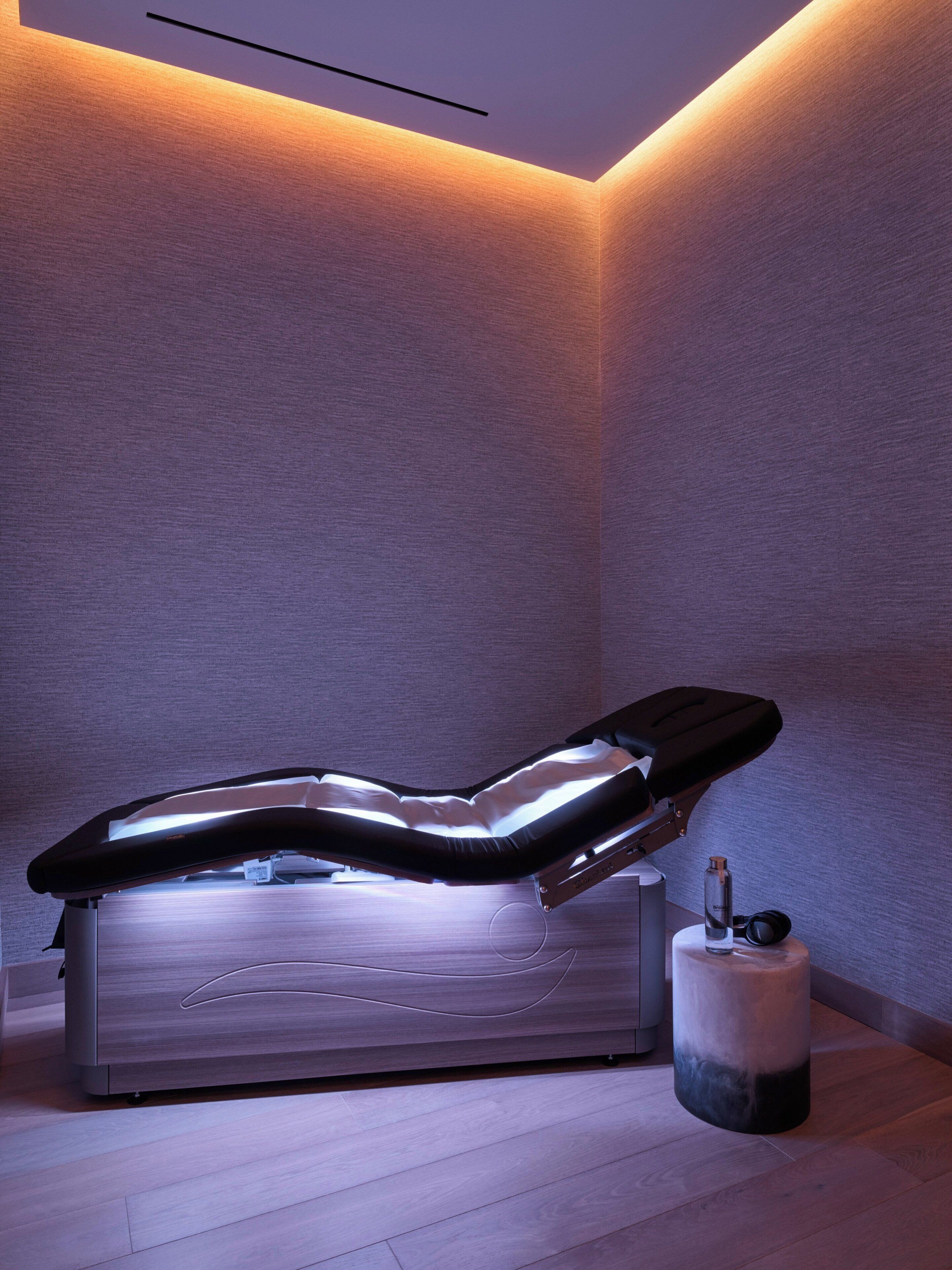 Un cuarto de terapia de masajes con una silla reclinable, una mesita con una botella de agua, y una lámpara fijada en la pared.