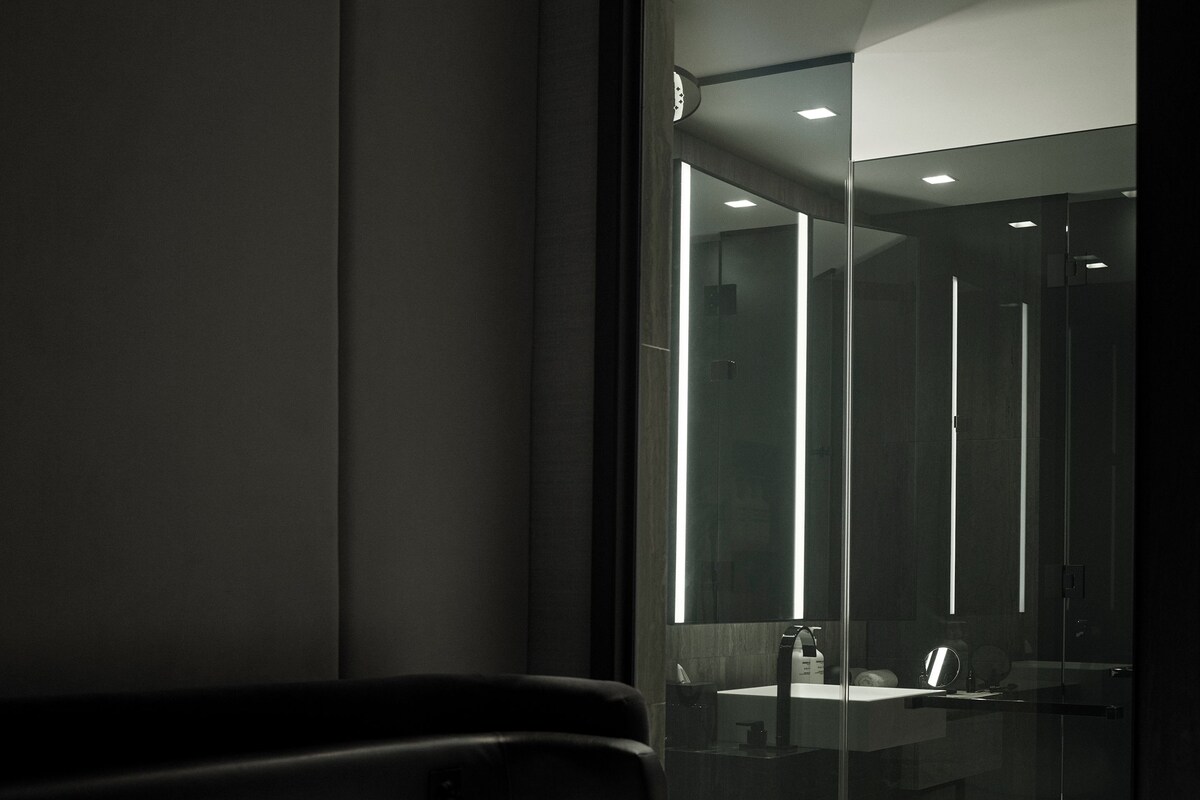 Une chambre d’hôtel moderne avec un lit, une cloison en verre et une salle de bain ouverte, équipée d’un lavabo et d’un miroir.