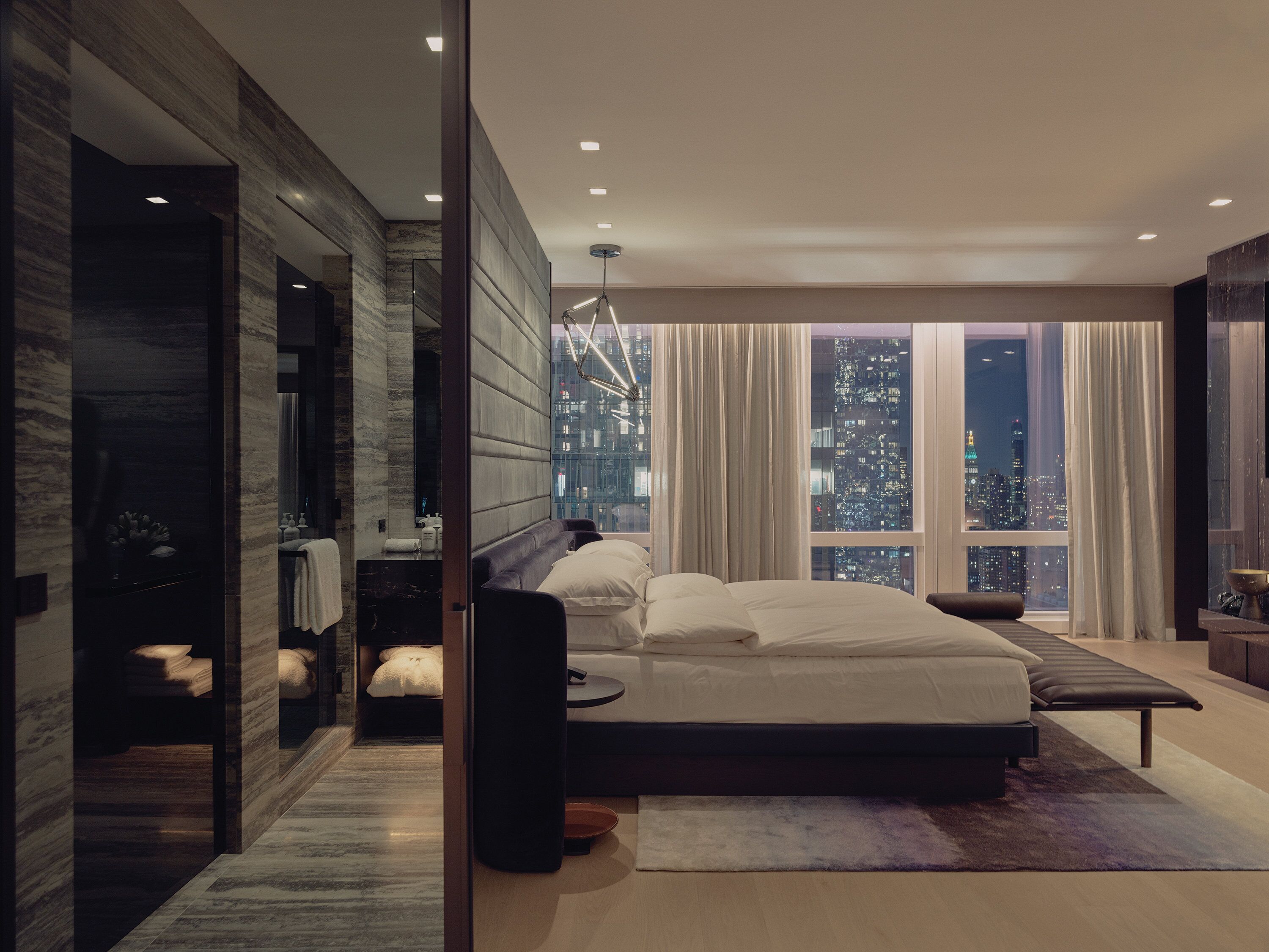 Ein modernes Schlafzimmer mit einem großen Bett, einer Sitzecke, einem Schminktisch und Blick auf die Stadt.