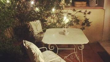 An outdoor patio with a table and two chairs, surrounded by greenery and string lights.