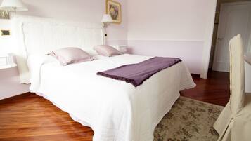 A neatly made bed with a white headboard, a purple throw blanket, and two pillows. There is a wooden floor and a floral-patterned rug.