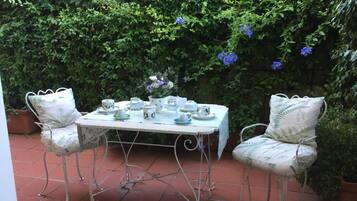 An outdoor patio with a table set for tea, surrounded by chairs and a lush green hedge in the background.
