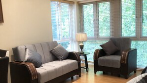 A modern living room with a grey sofa, a matching armchair, a small side table with a lamp, and large windows with blinds. - Park View 2 Bedroom By Creatick (Maidenhead)