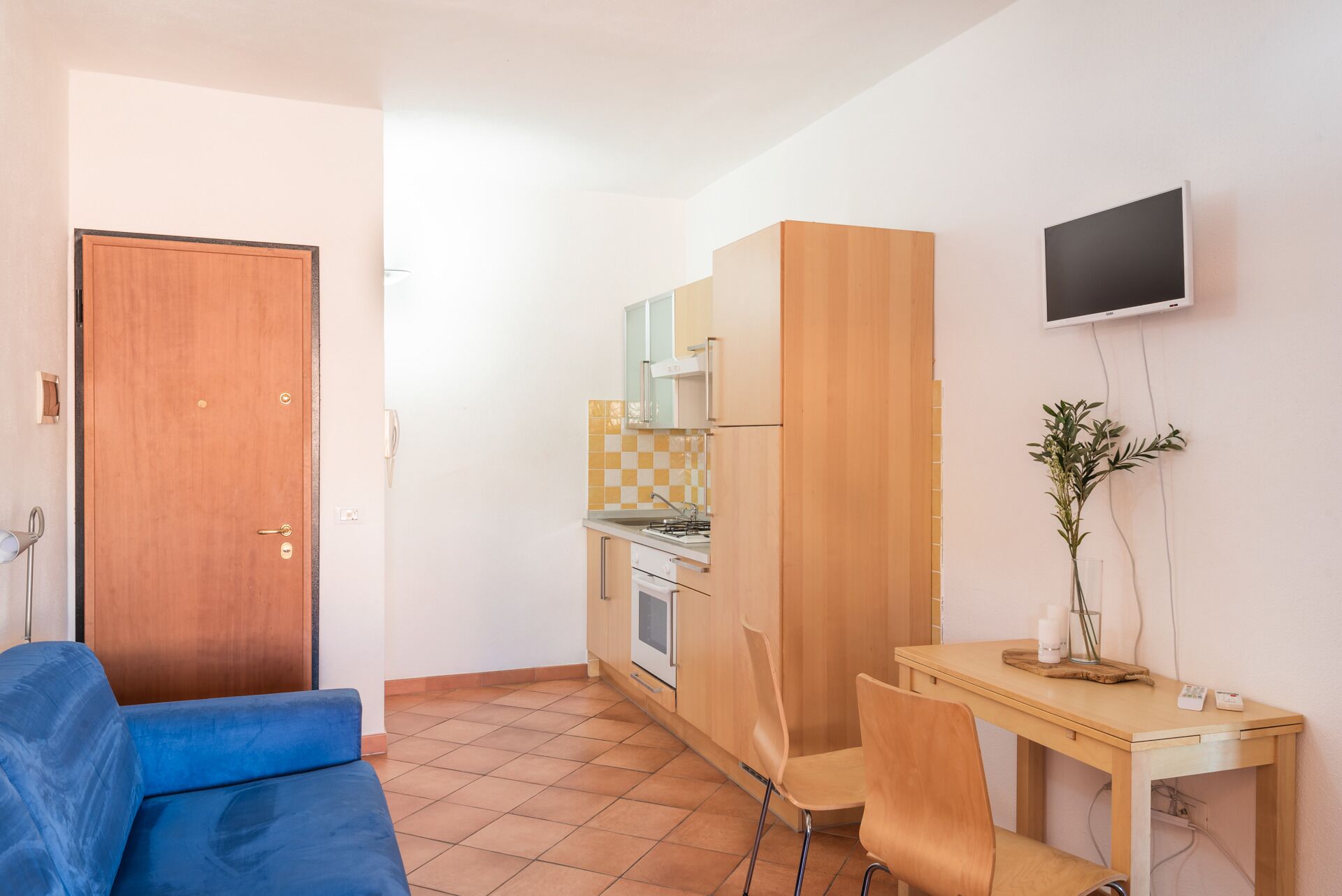 A compact kitchen with a stove, oven, and microwave. There is a wooden dining table with chairs and a small plant on the table. A blue sofa is positioned in the foreground.