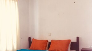 Uma cama de solteiro com colcha azul e duas almofadas laranja, uma cabeceira de madeira, um criado-mudo de madeira e uma parede branca com tomada.