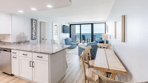 Una cocina moderna con armarios blancos, encimera de granito y una zona de comedor con mesa y sillas de madera.