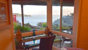 A room with a large window overlooking a body of water and hills. There is a glass table with a chair and a bed with a pillow.