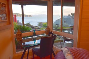 A room with a large window overlooking a body of water and hills. There is a glass table with a chair and a bed with a pillow.