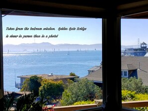 A view from a bedroom window overlooking the Golden Gate Bridge and the bay, with sailboats and buildings in the distance.