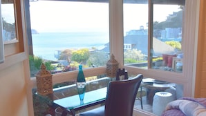 A room with a large bay window overlooking a scenic view, a small table with a chair, and a bed with a purple blanket.