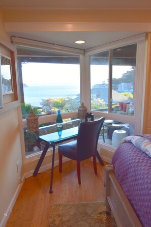 A room with a large bay window overlooking a scenic view, a small table with a chair, and a bed with a purple blanket.