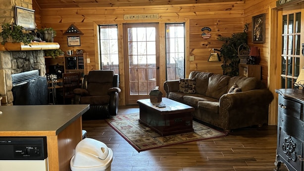 Un acogedor interior de cabaña con techo de madera, chimenea de piedra, comedor, sala de estar con sofá y mesa de centro, y área de cocina con lavavajillas.