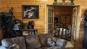 Un interior de cabaña acogedor con un sofá de cuero, una mesa de centro de madera, un cuadro enmarcado y un cartel que dice "CABIN SWEET CABIN".