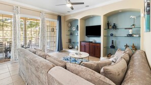 A living room with a beige sofa, a coffee table, and a TV. There are shelves with decorative items and a ceiling fan. - Reunion Views | Movie Room, Amazing Views of the Tom Watson Golf Course (Kissimmee)