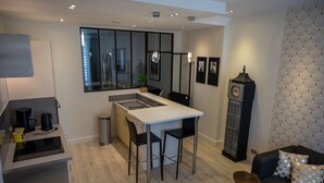 A compact kitchen with a central island, a dining area with two chairs, a sofa with patterned cushions, and a decorative clock.