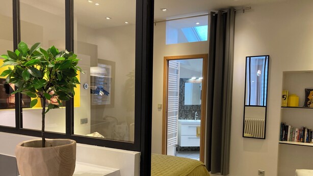 A modern bedroom with a large window, a bedside table, a bed with a green bedspread, a bedside lamp, and a bookshelf.
