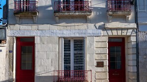A two-story building with a red door and windows, a balcony with a metal railing, and a chimney.