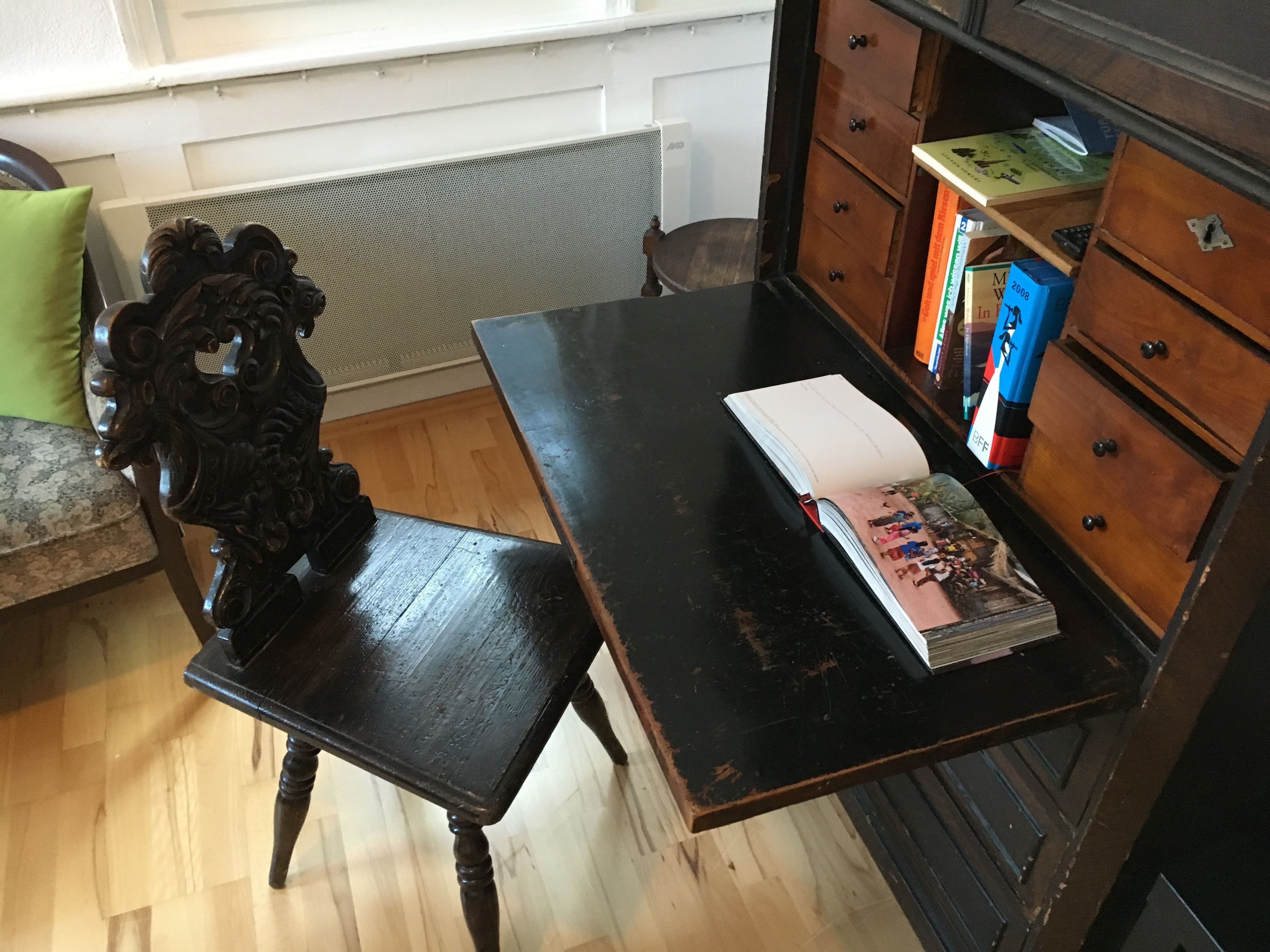 Una habitación con un escritorio de madera, una silla, un radiador y una estantería con libros y una caja.