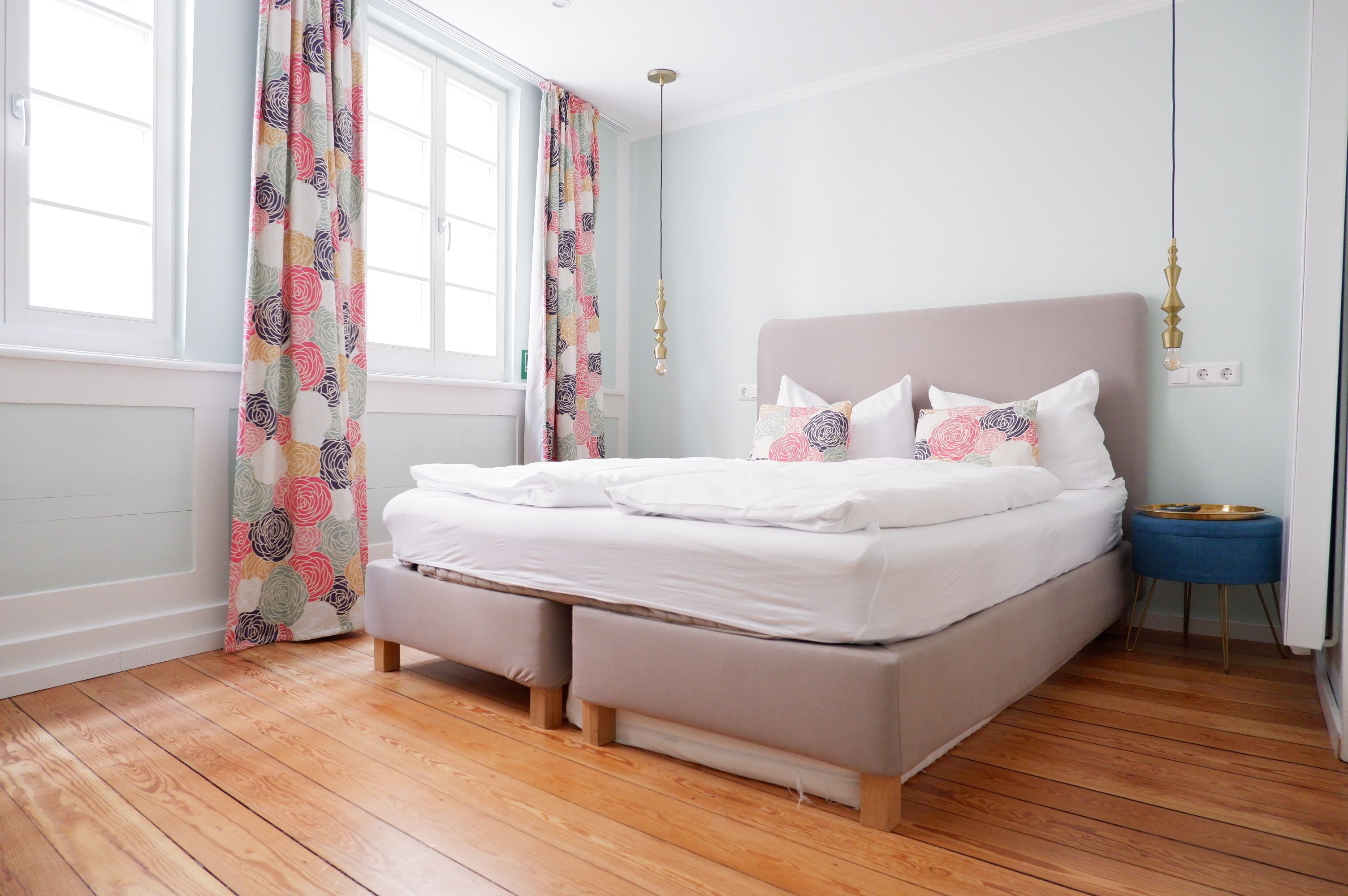 Un lit bien fait, avec du linge de lit blanc, un plancher en bois et une fenêtre ornée de rideaux à motifs floraux.