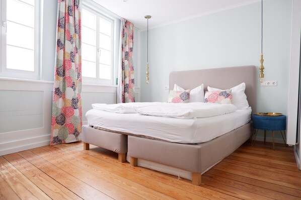 Una cama bien hecha con sábanas blancas, piso de madera y ventana con cortinas florales.