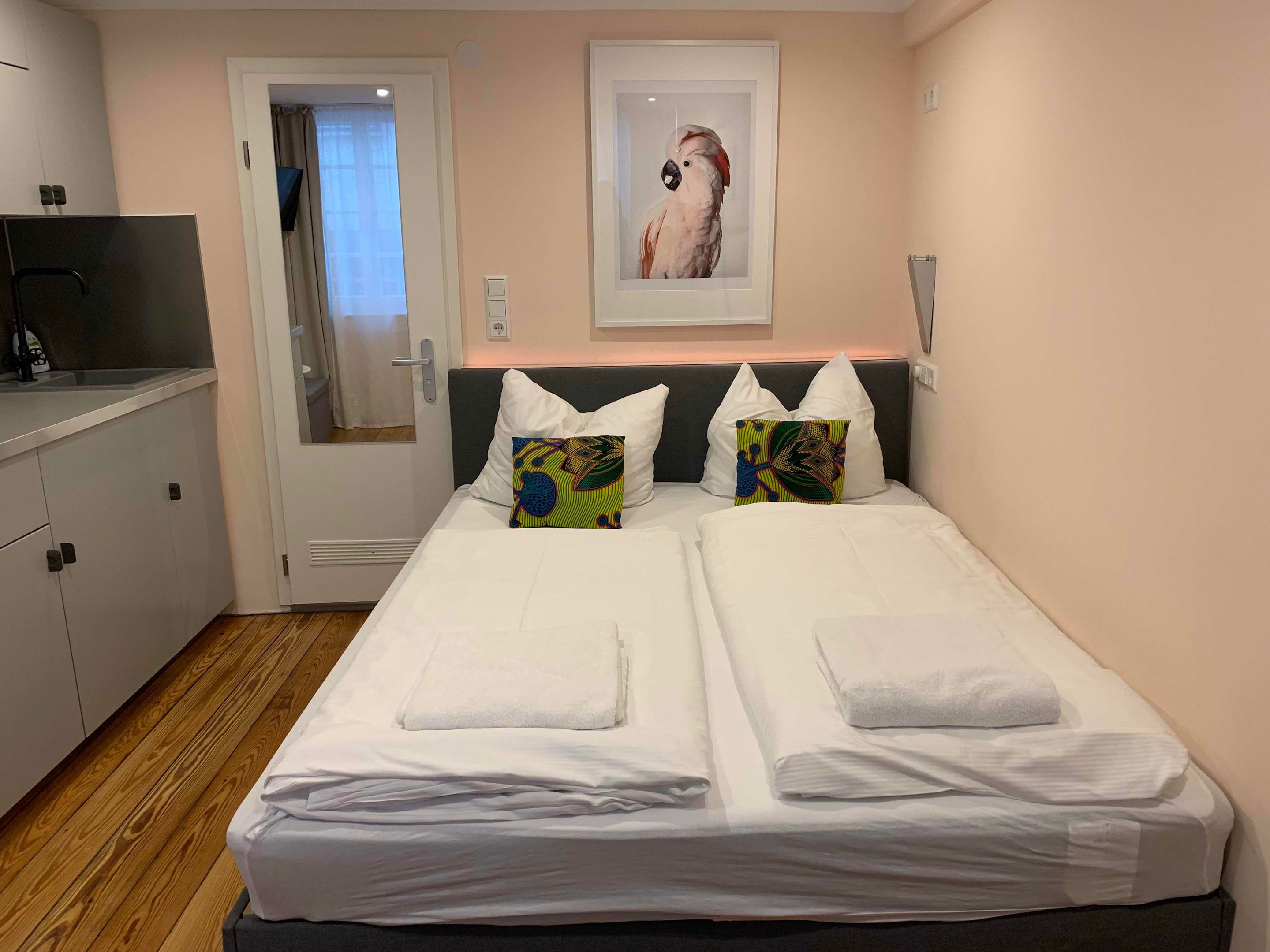 Quarto de hotel com cama, duas almofadas, um quadro emoldurado, uma escrivaninha e uma porta.
