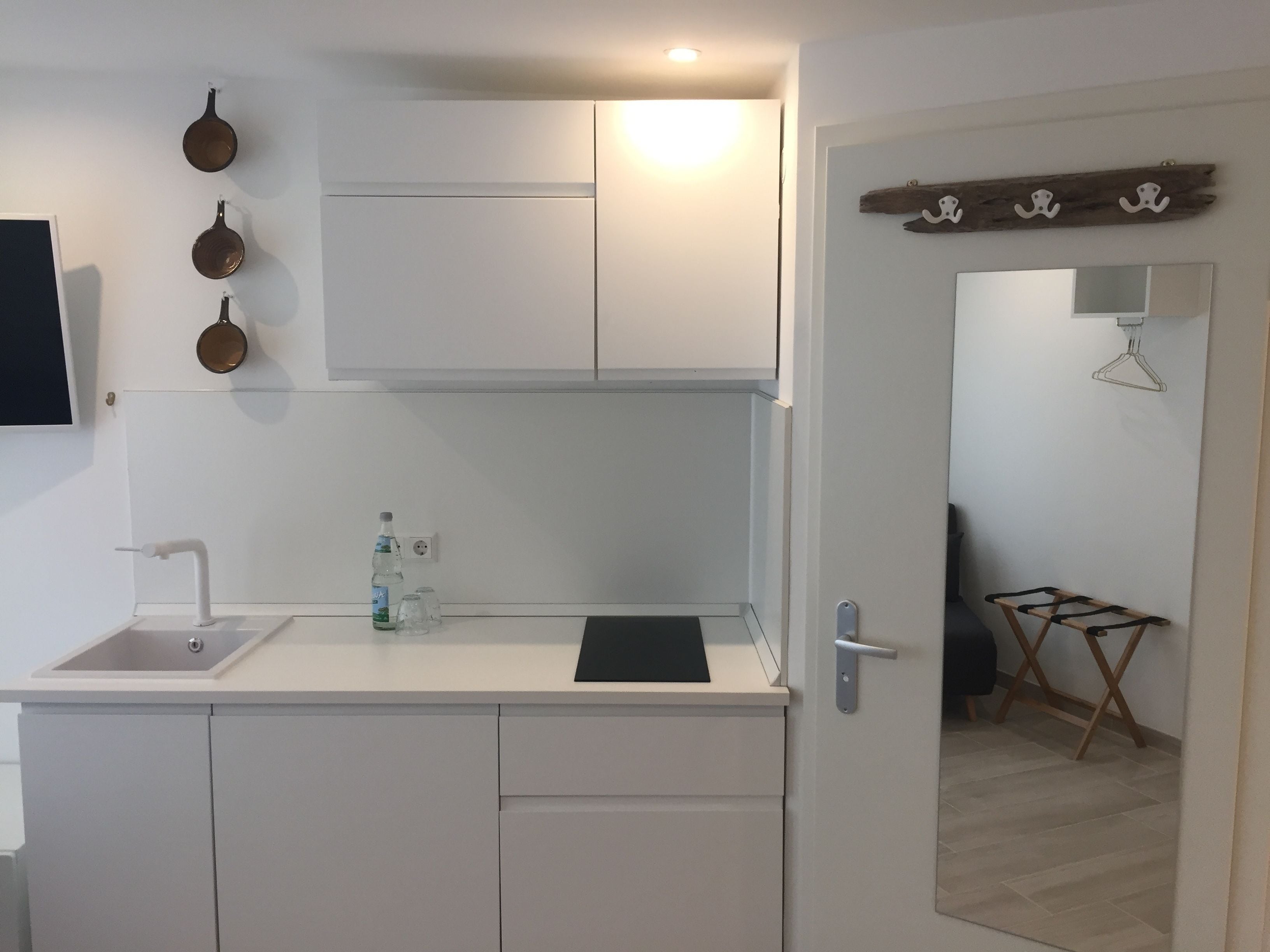 Una cucina compatta con un piano di lavoro bianco, un lavandino e una piccola finestra. Si vedono tre pentole appese al muro e un porta asciugamani di legno.