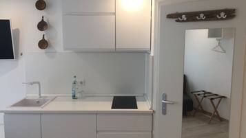 Una cucina compatta con un piano di lavoro bianco, un lavandino e una piccola finestra. Si vedono tre pentole appese al muro e un porta asciugamani di legno.