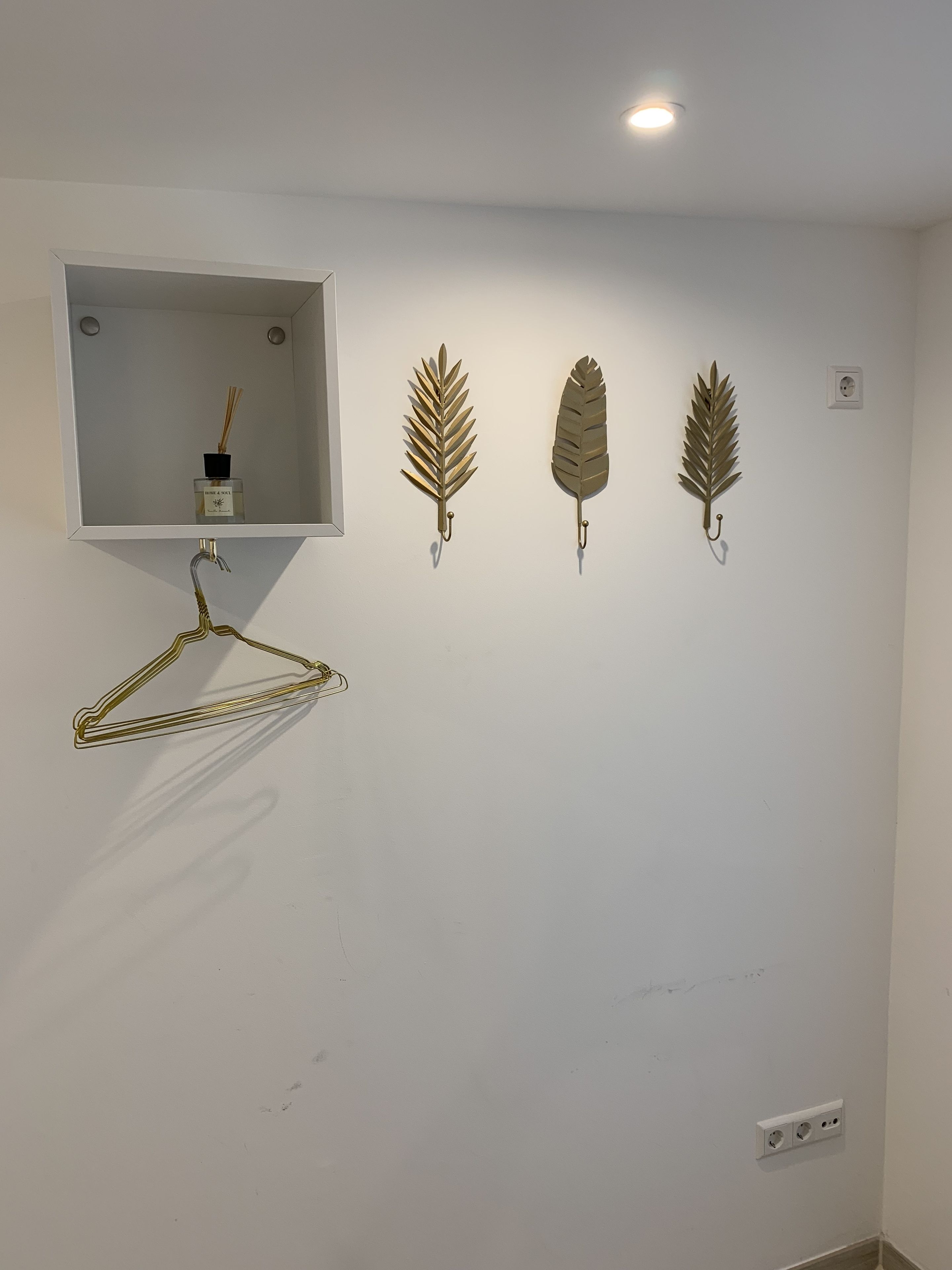 A wall with a white shelf holding a bottle of reed diffuser and four wall-mounted hooks with decorative items.