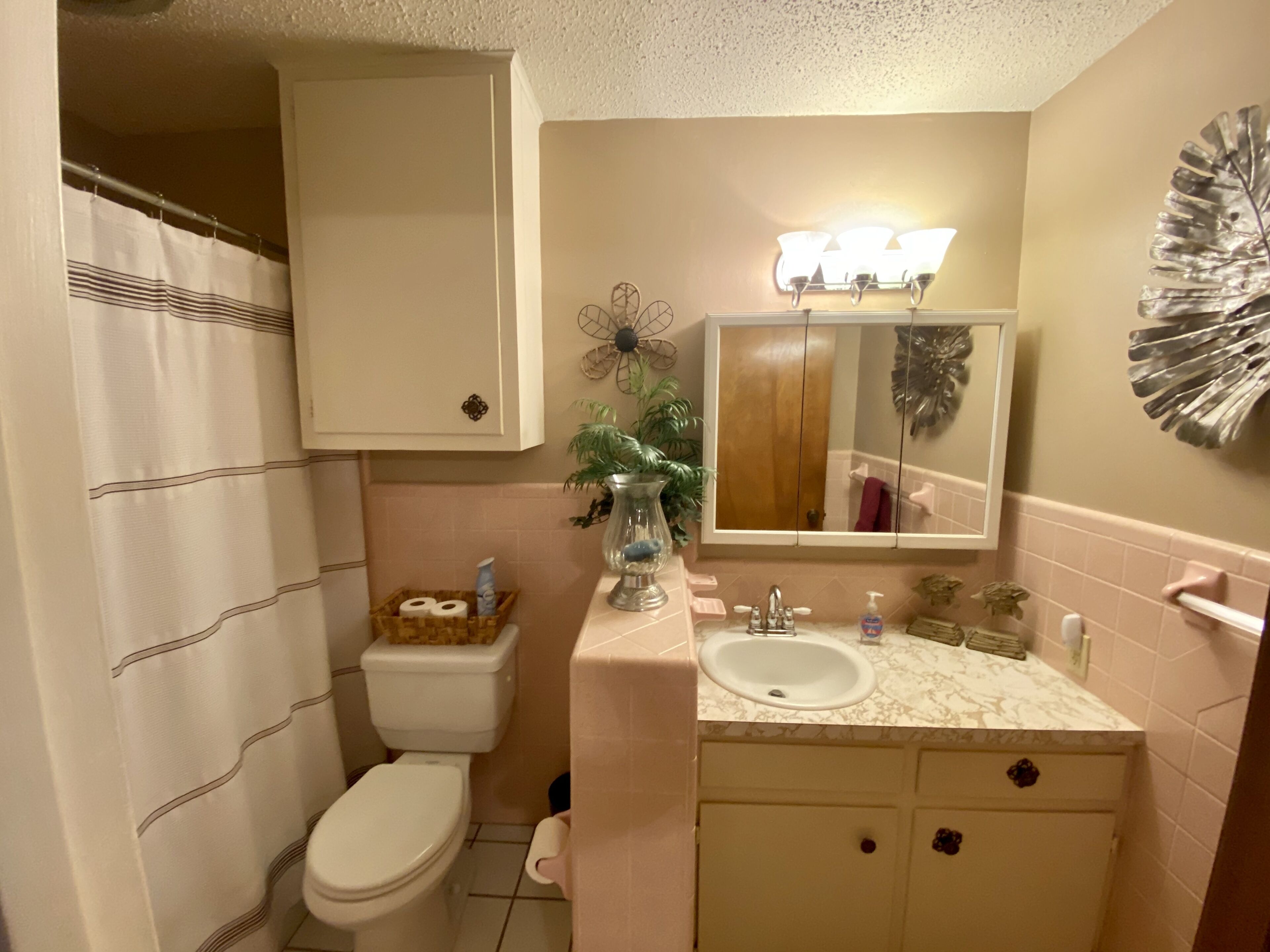 A bathroom with a toilet, sink, and mirror. There is a shower curtain, a vase with a plant, and a decorative mirror above the sink.