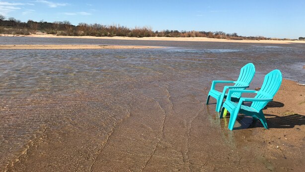 A beach with two turquoise chairs and a table, a sandy shore, and a clear sky.
