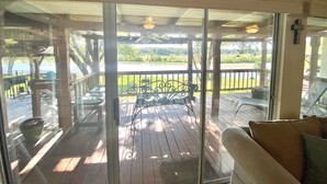 A covered outdoor area with a wooden deck, a metal bench, and a glass door leading to a view of a lake and greenery.