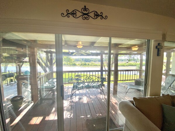 A covered outdoor area with a wooden deck, a metal bench, and a glass door leading to a view of a lake and greenery.