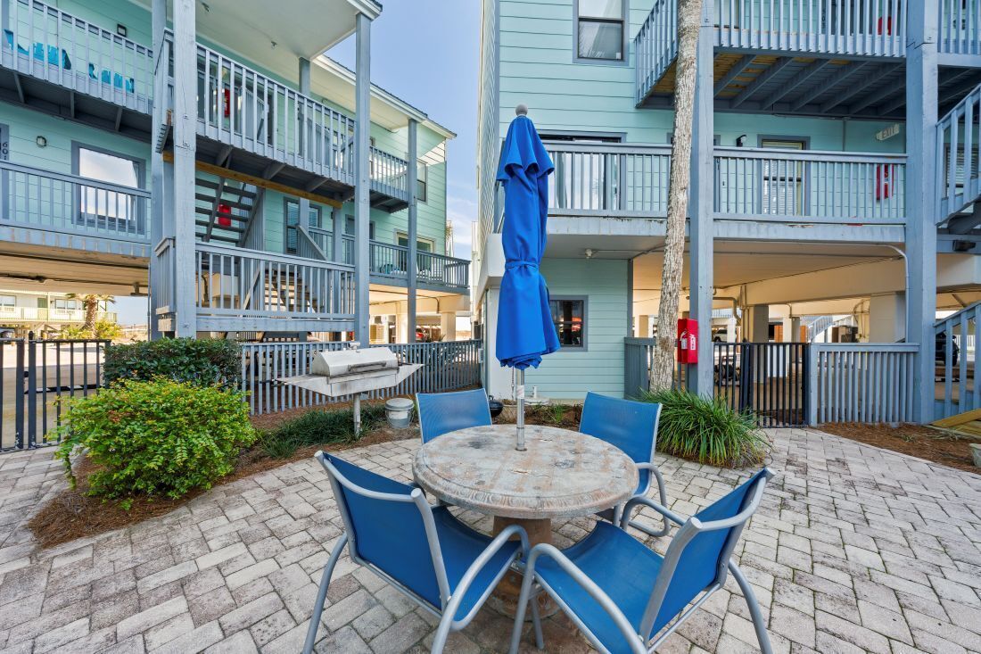 A patio with a round table and blue chairs, surrounded by a brick walkway and greenery.
