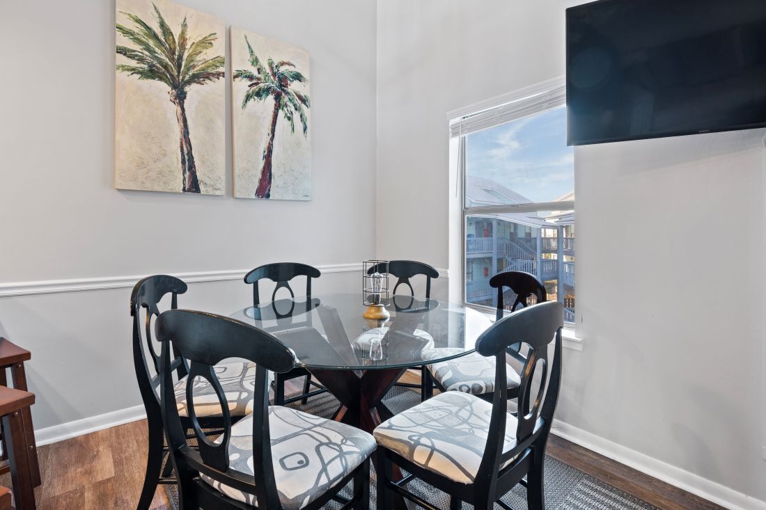 Un coin repas avec une table en verre, des chaises noires et deux tableaux représentant des palmiers accrochés au mur.