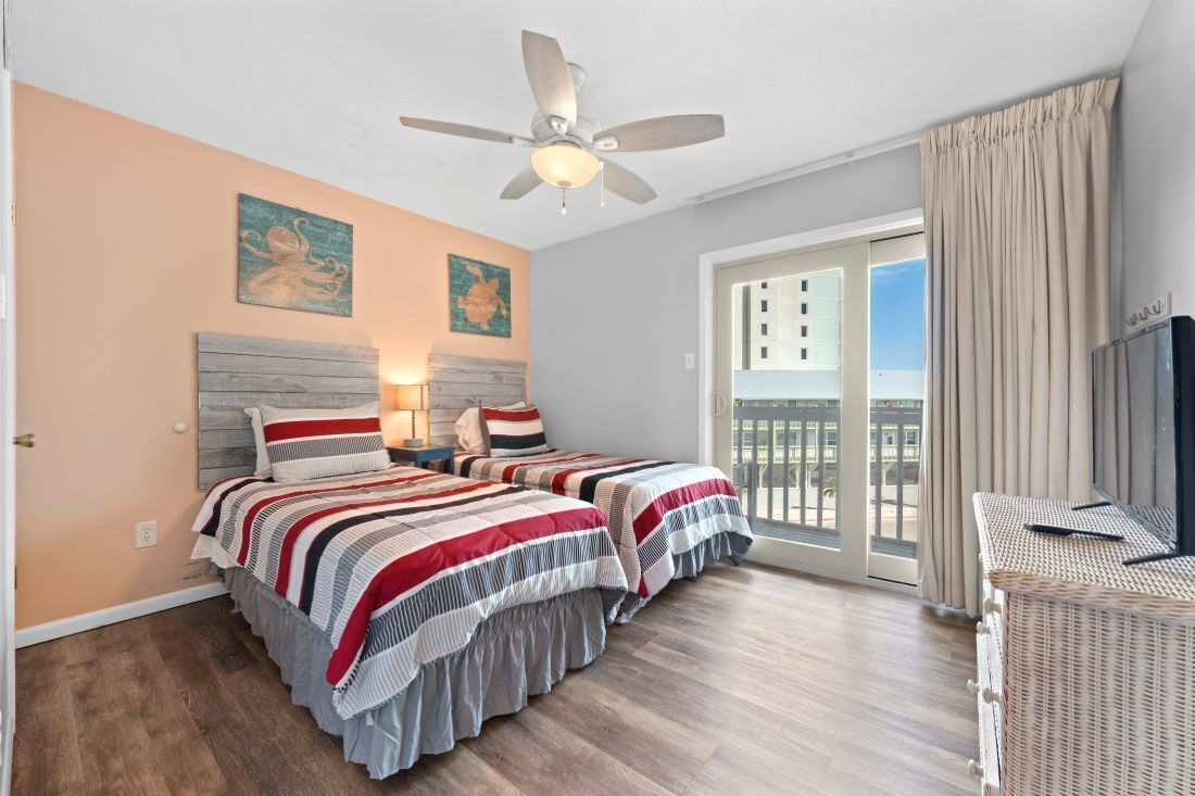 Une chambre avec deux lits, un ventilateur de plafond, un balcon avec des rideaux et des œuvres d’art accrochées aux murs.