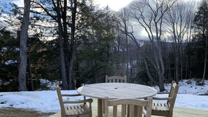 Een houten terras met een ronde tafel en vier stoelen, omringd door een met sneeuw bedekte grond en bomen.