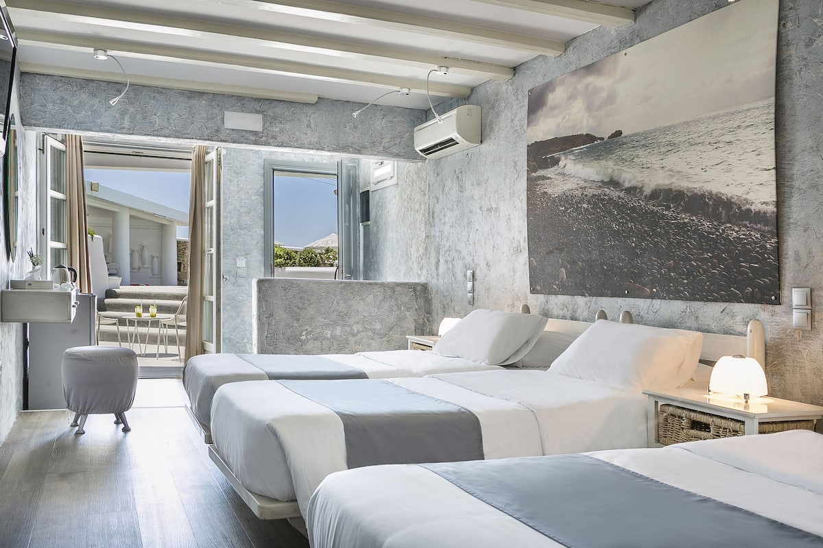 A modern hotel room with two beds, a large painting of a beach, and a view of the outdoors.