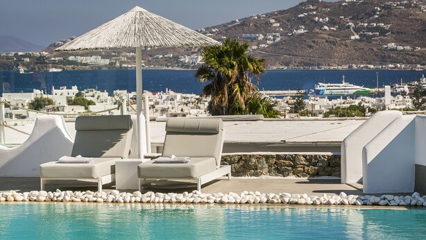 Área de piscina com espreguiçadeiras e um guarda-sol de palha, com vista para uma vila costeira e o mar.